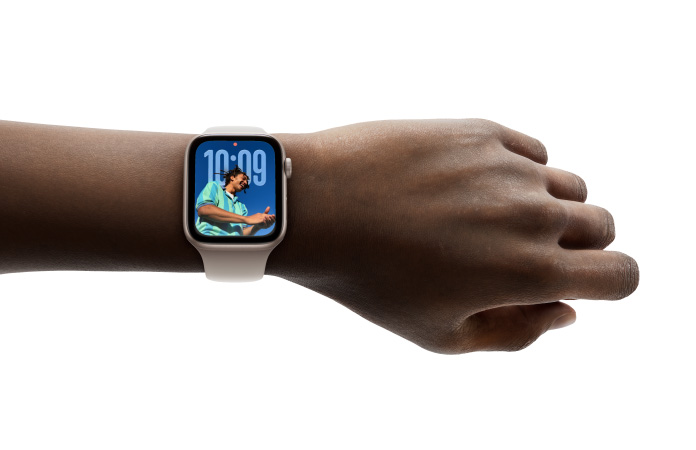 Bir kişinin bileğindeki kauçuk kordonlu Gümüş rengi Apple Watch’un yakından çekilmiş görseli, kadranda yeni Liquid Glass tasarımıyla Saat uygulaması görülüyor
