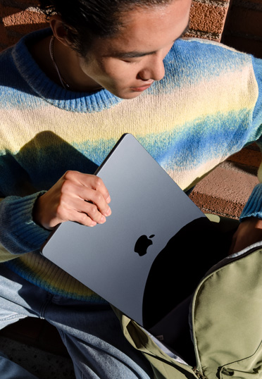 一名學生坐在戶外。他從背囊中拿出一台合上的 MacBook。